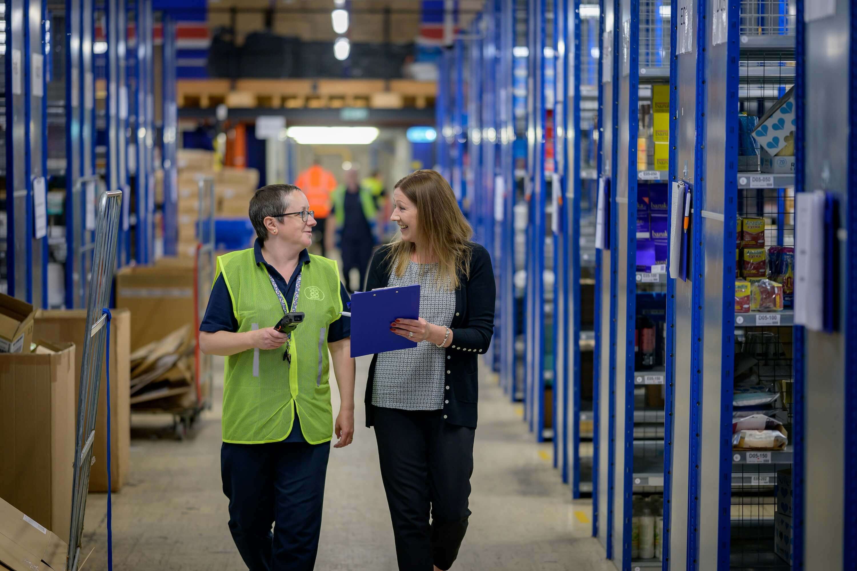 Two people walking through a warehouse aisle, holding a handheld scanner and a clipboard while doing an inventory.