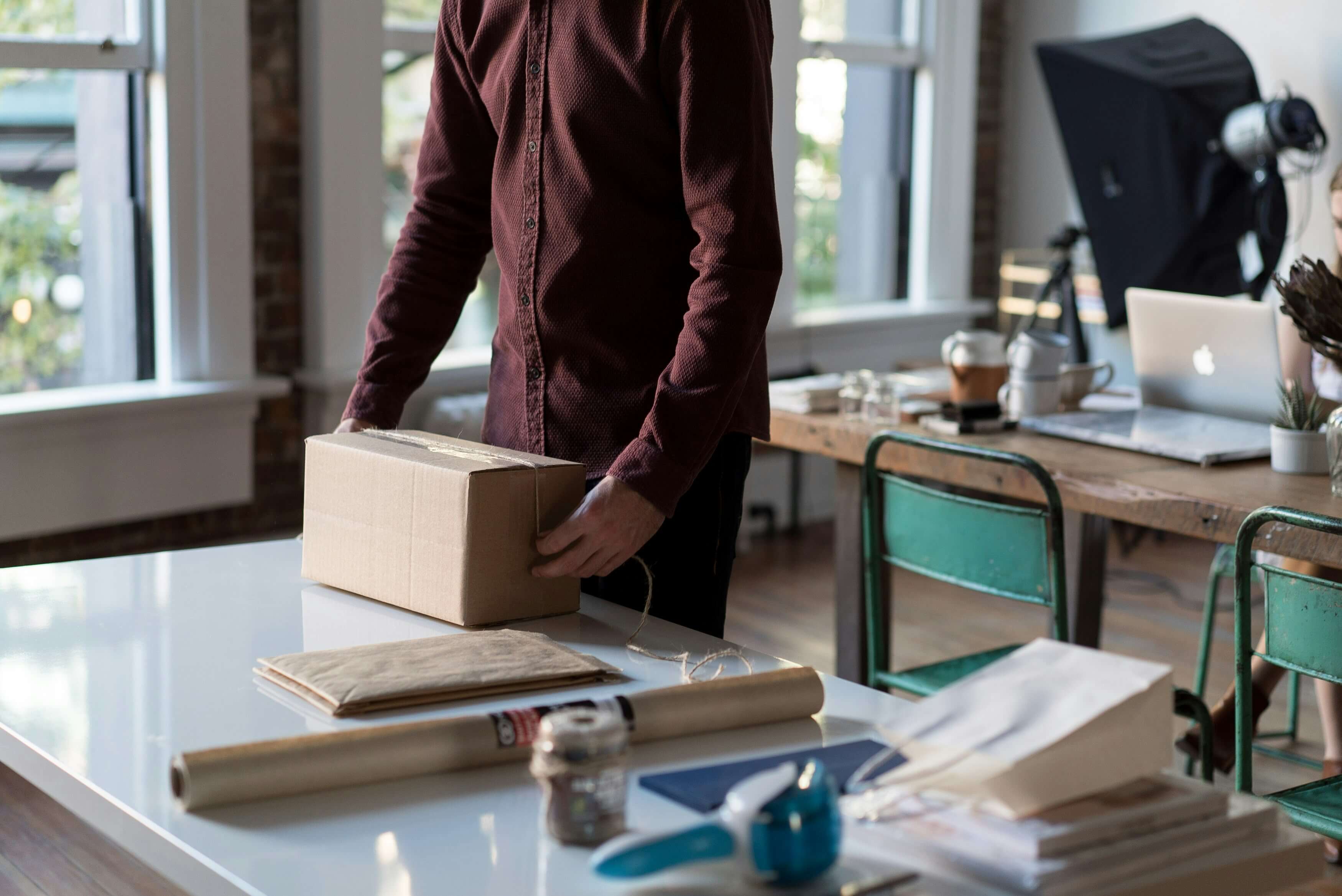 Person holding a cardboard box in a workspace with desks and various items.