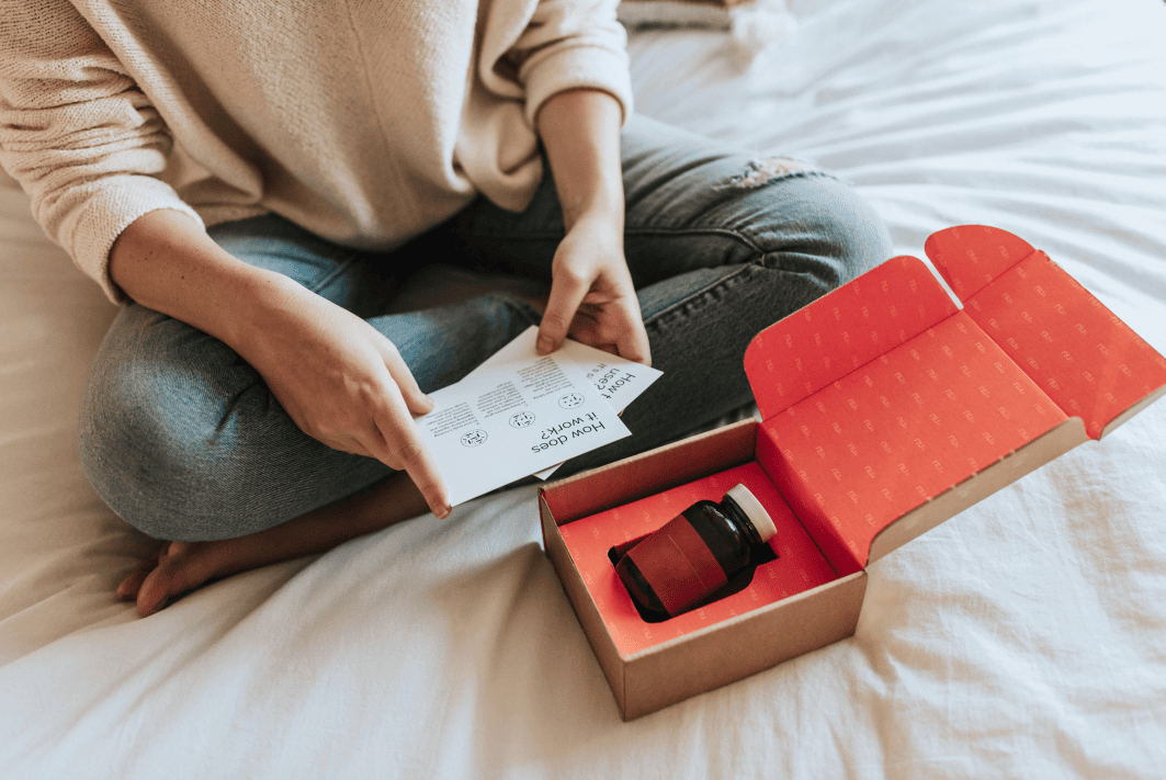 Personne assise sur un lit, tenant une feuille de papier à côté d'une boîte en carton ouverte contenant une bouteille foncée avec un bouchon blanc et une étiquette rose.